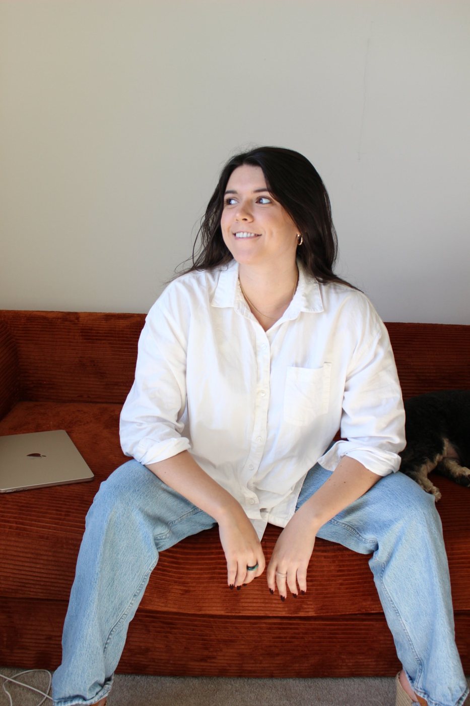 Elizabeth laughing and looking off to the side, sitting on a sofa in a white shirt and jeans