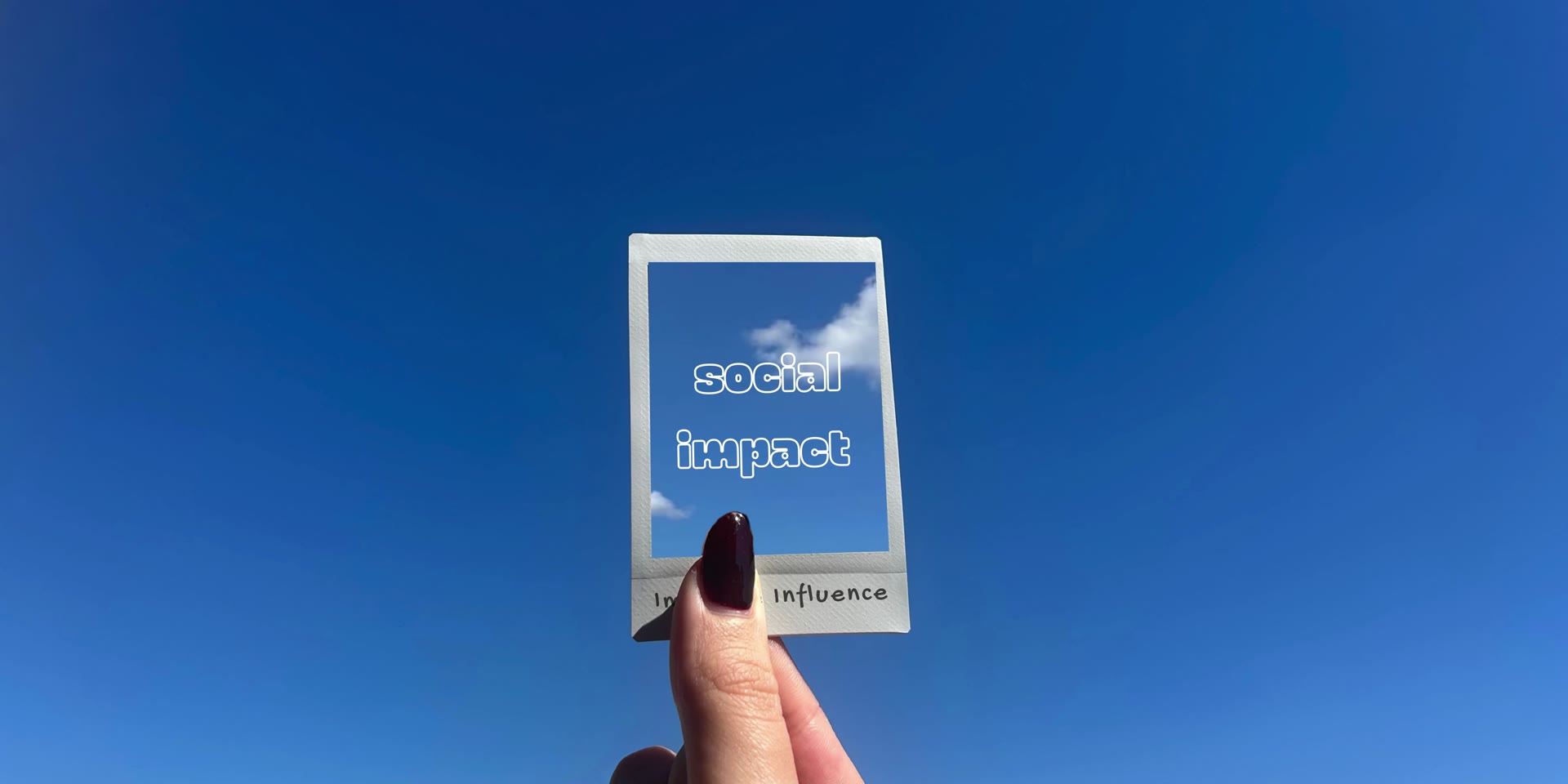 A hand holding a polaroid that reads social impact against a blue sky